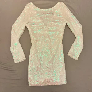 Paljettklänning - Snygg klänning täckt av glittrande paljetter som ger en skimrande effekt. Klänningen har en v-ringad design i rugged och långa ärmar, perfekt för en festlig kväll. Den är kort och figurnära, vilket ger en elegant silhuett.