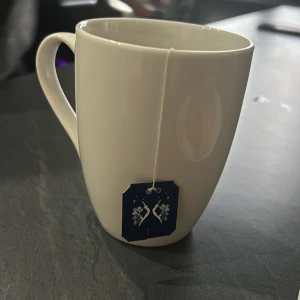 Mugg - Vit keramikmugg med ett te-påse hänge. Muggen har ett klassiskt handtag och en slät yta, perfekt för varma drycker som te eller kaffe. Enkel och stilren design som passar i alla kök.