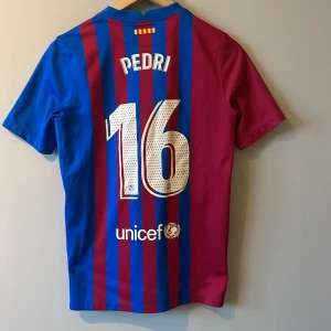 Blåröd fotbollströja från Nike - Säljer en blåröd randig fotbollströja från Nike med korta ärmar. Tröjan har tryck med 'Pedri' och nummer 16 på ryggen samt La Liga-märke på ärmen. Perfekt för fans av FC Barcelona.