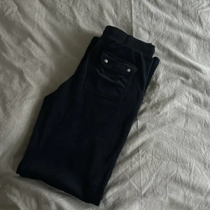 Marinblå mjukisbyxor från Juicy Couture - Säljer ett par marinblå  mjukisbyxor från Juicy Couture med snörning i midjan och en broderad logga. Byxorna har en ficka med knappar på sidan och är perfekta för en avslappnad stil.
