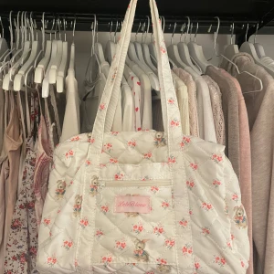 Blommig quiltad väska coquette - Charmig quiltad väska med blommigt mönster och söta nallemotiv. Väskan har en praktisk dragkedja med hjärtformad detalj och en rosa etikett framtill. Perfekt för att ge en söt touch till din outfit. Den har massa fack och det får plats mycket. 32cm lång, 13cm bred, 22cm hög. Handtagen; 23cm långa