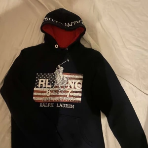 Unik Ralph Lauren hoodie - En riktigt fet unik mörkblå hoodie från Ralph Lauren med en stor logga på framsidan. Hoodien har en röd insida i huvan och texten 'Ralph Lauren' på kragen. Perfekt för en avslappnad stil. Den är i väldigt bra skick skriv vid minsta fråga eller fundering💯👍