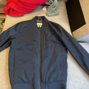 Mörkblå bomberjacka från Hilfiger Denim - Snygg mörkblå bomberjacka från Hilfiger Denim med dragkedja framtill och en bröstficka med dragkedja. Jackan har en klassisk bomberstil med ribbade muddar vid ärmslut och nederkant. 