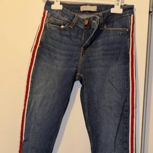 Jeans med ränder från Zara - Snygga jeans från Zara med röda och vita ränder längs sidorna. De har en klassisk femficksdesign och knappgylf. Perfekta för en trendig look!
