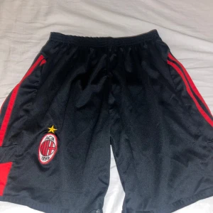 Svarta fotbollsshorts med röd detalj - Snygga svarta fotbollsshorts med röda detaljer och AC Milan-logga. Perfekta för fotbollsträningen eller matchen. Elastisk midja för bekväm passform. Storleken finns inte men passar XS-S