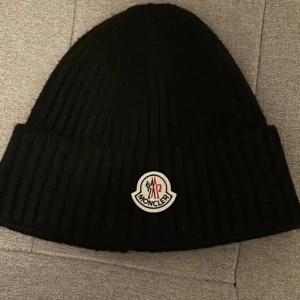 Svart ribbad mössa från Moncler - Snygg svart ribbad mössa från Moncler med broderad logotyp framtill. Perfekt för att hålla värmen under kyliga dagar.