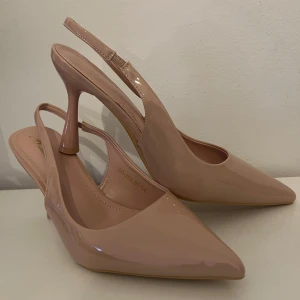 Pummpsu - Eleganta beige pumps med spetsig tå och slingback-design. Skorna har en hög klack och är tillverkade i ett glansigt material som ger en stilren look.