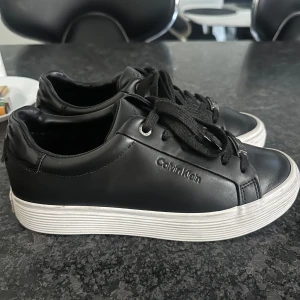 Svarta sneakers från Calvin Klein - Snygga svarta sneakers från Calvin Klein i skinn med snörning. Skorna har en vit sula och diskret logotyp på sidan och hälen. Perfekta för en stilren look.