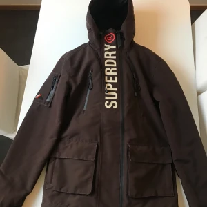 Brun jacka från Superdry - Snygg brun jacka från Superdry med dragkedja och stora fickor. Jackan har en hög krage och är perfekt för kyligare dagar. Den har en tydlig logga på framsidan och en huva för extra skydd. Använt 3 gånger så skick är som nya ! Storlek S