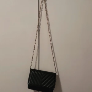 Svart quiltad axelväska med kedja - Snygg svart axelväska med quiltat mönster och guldfärgad kedja. Perfekt för att ge en stilren touch till din outfit. Väskan har en praktisk knappstängning och är tillverkad i syntetmaterial.