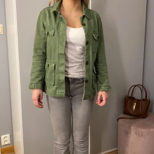 Grön jeans jacka med knappar - Säljer en snygg grön jacka med knappar framtill och två stora fickor. Jackan har en avslappnad passform och långa ärmar. Perfekt för vår och höst!