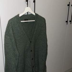Säljer en stilren grön kofta från Massimo Dutti. Den är stickad med en v-ringad design och har knappar framtill. Perfekt för att hålla sig varm och snygg under kyligare dagar.