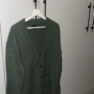 Grön kofta från Massimo Dutti - Säljer en stilren grön kofta från Massimo Dutti. Den är stickad med en v-ringad design och har knappar framtill. Perfekt för att hålla sig varm och snygg under kyligare dagar.