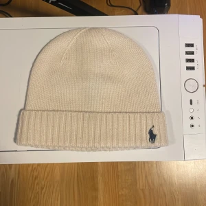 Beige stickad mössa från Ralph Lauren - Snygg beige stickad mössa från Ralph Lauren med broderad logga i mörkblått. Perfekt för kyliga dagar med sin ribbade kant som ger extra värme och stil.               Ny pris 699kr