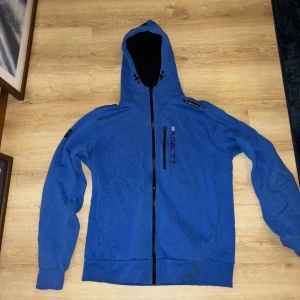 Blå hoodie med dragkedja - Säljer en blå hoodie med dragkedja och tryck på bröstet. Den har en justerbar huva och långa ärmar. Perfekt för en avslappnad stil.