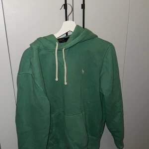 Grön hoodie från Ralph Lauren - Säljer en snygg grön hoodie från Ralph Lauren med klassisk känguruficka och justerbar dragsko i huvan. Perfekt för en avslappnad stil. Den har en liten logotyp broderad på bröstet.