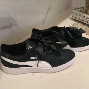 Svarta sneakers från Puma med satinband - Snygga svarta sneakers från Puma med vita detaljer och unika satinband som snörning. Perfekta för en stilren look med en twist. Puma-loggan syns tydligt på sidan och tungan av skon.