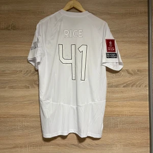 Vit Arsenal fotbollströja med nummer 41 - Säljer en vit Arsenal fotbollströja från Adidas med nummer 41 och namnet 'RICE' på ryggen. Tröjan har korta ärmar och FA Cup-märke på ärmen. Perfekt för fans som vill visa sitt stöd.
