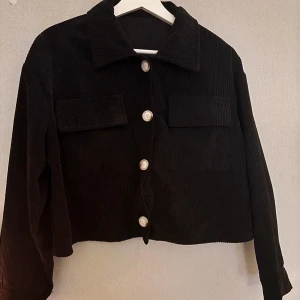 Svart manchesterjacka med knappar - Snygg svart manchesterjacka med stora knappar framtill och två bröstfickor. Jackan har en croppad design och långa ärmar, perfekt för en trendig look. Passar bra till både jeans och kjol.