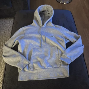 Grå hoodie från Lacoste - Säljer en stilren grå hoodie från Lacoste med klassisk passform. Tröjan har en känguruficka och den ikoniska krokodilloggan på bröstet. Perfekt för en avslappnad look.