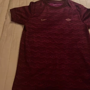 Vinröd fotbollströja från Nike - Snygg vinröd fotbollströja från Nike med ett geometriskt mönster och guldiga detaljer. Tröjan är kortärmad och tillverkad i Dri-FIT-material för optimal komfort. Perfekt för både träning och match!