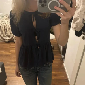 Mörkblå blus med knytning - Säljer en söt mörkblå blus med korta puffärmar och knytning framtill. Blusen har en volangdetalj vid midjan som ger en fin siluett. Perfekt för en stilren look.