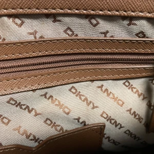 Bordeaux  och beige-färgad  handväska från DKNY - Snygg handväska från DKNY i bordeux och beige med monogrammönster. Väskan har en stilren design med metallspänne framtill och dubbla handtag. Insidan är fodrad med DKNY-logga och har en praktisk dragkedjeficka.