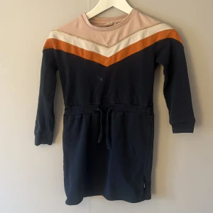 Flerfärgad klänning från Minymo - Säljer en cool klänning från Minymo i storlek 128. Den har ett snyggt mönster med mörkblå, orange och beige färger samt en glittrig detalj. Klänningen har långa ärmar och ett knytband i midjan för en perfekt passform. Perfekt för höst och vår!