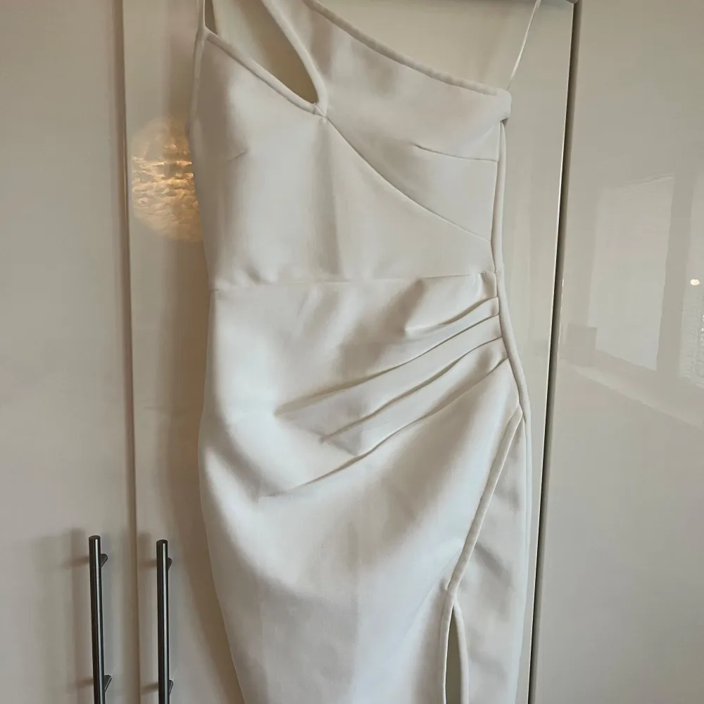 Elegant vit klänning med en stilren one-shoulder design och smickrande passform. Klänningen har en slits på sidan och asymmetriska detaljer som ger en modern touch. Perfekt för speciella tillfällen.. Mekot.