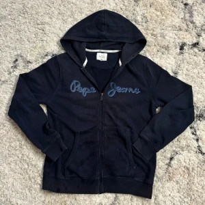 Marinblå hoodie från Pepe Jeans - Snygg marinblå hoodie från Pepe Jeans med dragkedja och huva. Den har ett stilrent tryck på bröstet och praktiska fickor framtill. Perfekt för en avslappnad stil.