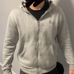 Grå hoodie från Burberry - Säljer en stilren grå hoodie från Burberry med dragkedja och fickor framtill. Perfekt för en avslappnad look. Den har en klassisk design med en liten logotyp på bröstet. Kan trycka in snöre på hoodien om ni vill och pris kan diskuteras 