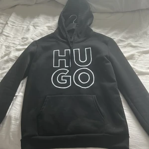 Svart hoodie från Hugo - Säljer en stilren svart hoodie från Hugo med stor vit logga på framsidan. Tröjan har en klassisk känguruficka och justerbar huva med dragsko. Perfekt för en avslappnad look.