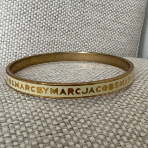 Guldarmband från Marc by Marc Jacobs - Elegant guldarmband från Marc by Marc Jacobs med en stilren design. Armbandet har märkets namn ingraverat runt om och en slät yta som ger en lyxig känsla. Perfekt för att lägga till en touch av glamour till din outfit. 
