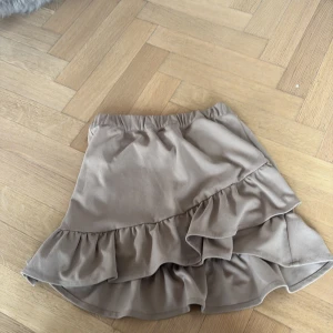 Beige kjol med volang - Säljer en söt beige kjol med volangdetaljer. Kjolen har en omlott-design och är perfekt till sommaren. Den är kort och har en elastisk midja för bekväm passform.💕