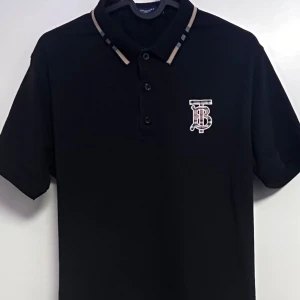 Svart pikétröja med logga - Snygg svart pikétröja från Burberry med korta ärmar och klassisk krage. Tröjan har en stilren logga på bröstet och knappar framtill. Perfekt för en avslappnad men ändå stilren look.