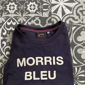 Marinblå tröja från Morris - Säljer en stilren marinblå tröja från Morris med texten 'MORRIS BLEU MARINE' på framsidan. Tröjan har långa ärmar och en ribbad krage med röd och vit randig detalj. Perfekt för en avslappnad look.