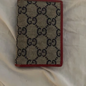 Gucci Mönstrad plånbok med röd kant - Snygg plånbok med ett klassiskt mönster och röd kant. Insidan är röd med flera fack för kort och en genomskinlig sektion. Perfekt för att hålla ordning på dina kort och kontanter.