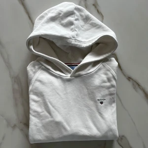 Vit hoodie från GANT - Säljer en stilren vit hoodie från GANT med broderad logga på bröstet. Tröjan är varm och skön inuti och snygg från utsidan. Passar bra till både sommarkvällar samt resten av året. Perfekt för en avslappnad look.