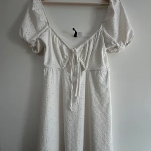 Vit klänning med puffärmar - Charmig vit klänning med puffiga kortärmar och knytning framtill. Klänningen har ett vackert broderat mönster och en luftig passform som ger en romantisk känsla. Perfekt för sommarens alla tillfällen men även studenten