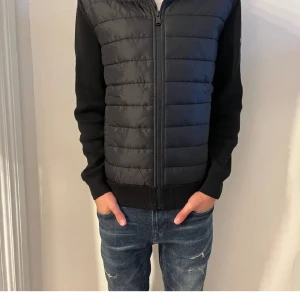 Svart jacka från Barbour - Snygg svart jacka från Barbour med quiltad framsida och dragkedja. Jackan har långa ärmar och en huva baktill. Perfekt för en stilren look. Passar 160 - 165!