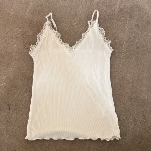 Vit ribbad linnetopp med spetsdetaljer - Säljer en stilren vit ribbad linnetopp med vackra spetsdetaljer längs v-ringningen och axelbanden. Perfekt för varma dagar eller som ett lager under en kavaj. Justerbara axelband för bästa passform.