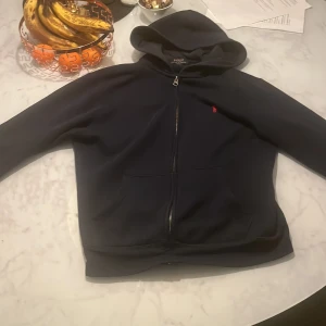 Mörkblå hoodie från Ralph Lauren - Säljer en snygg mörkblå hoodie från Ralph Lauren med dragkedja och huva. Den har en röd broderad logotyp på bröstet och praktiska fickor framtill. Perfekt för en avslappnad stil. Strl är xl barn men passar xs till s pris ej hugget i sten hör av er vid frågor!🍾😊