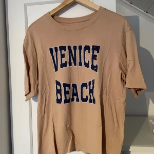 Beige t-shirt från Lager 157 - Säljer en beige t-shirt med texten 'Venice Beach' i mörkblått. Rund hals. Från lager 157. Storlek M