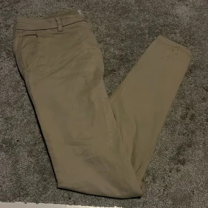 Snygga och stilrena somriga Lindbergh chinos - Snygga beige chinos från Lindbergh med en klassisk design. Byxorna har en knapp och dragkedja framtill samt hällor för bälte. Perfekta för en stilren look på sommaren. Storlek 32/30. Byxorna har en liten fläck längst ner på byxbenet och jag vet inte om det går bort i tvätten. Hör av er om ni har några frågor och priset är såklart inte hugget i sten