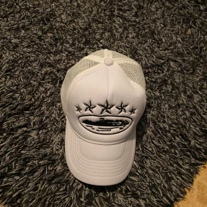 Vit keps med stjärnor - Snygg vit keps med broderade svarta stjärnor och ett coolt motiv framtill. Kepsen har ett justerbart spänne baktill och meshpaneler för extra ventilation. Perfekt för en avslappnad stil.
