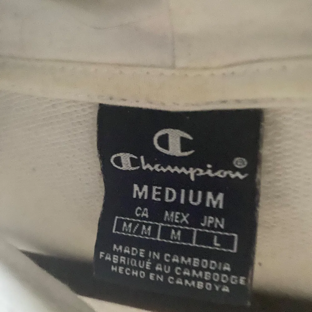 Vit hoodie från Champion med tryck i rött och blått på bröstet. Den har en klassisk känguruficka och Champion-logga på ärmen. . Neuletakit & Villapaidat.