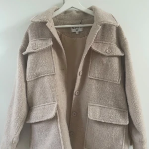 Beige kappa från NA-KD - Säljer en snygg beige kappa från NA-KD med stora fickor och knappar framtill. Den är i storlek 32 men är oversized så passar upp till 34.