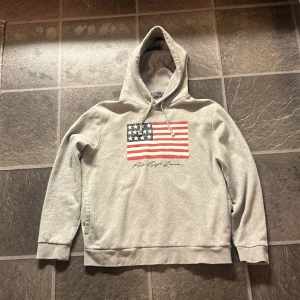 Grå hoodie från Polo Ralph Lauren - Säljer en grå hoodie från Polo Ralph Lauren med en stor amerikansk flagga på framsidan. Köpt i affär och den är knappt använd!