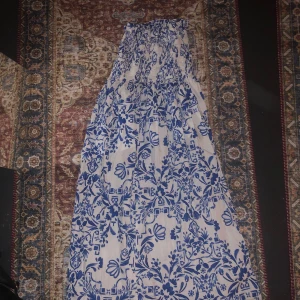 Vit och blå långkjol - Säljer en vacker vit långkjol med blått blommönster. Kjolen har en smockad överdel som ger en bekväm passform. Vet ej vart ifrån kjolen är. Perfekt för somriga dagar eller speciella tillfällen. Hör av dig vid intresse! 🌸