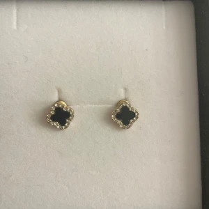 Svarta och guldiga örhängen med stenar - Eleganta örhängen i svart och guld med en fyrklöverformad design. De är dekorerade med gnistrande stenar runt kanterna som ger en lyxig känsla. Perfekta för att lägga till en touch av glamour till din look.